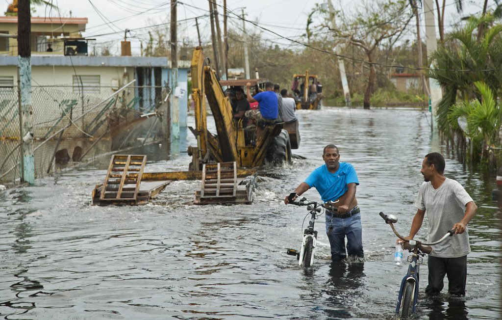 Two men walk their bicycles through floodwaters while recovery crews work in the background, following Hurricane Milton’s destruction.