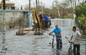 Two men walk their bicycles through floodwaters while recovery crews work in the background, following Hurricane Milton’s destruction.