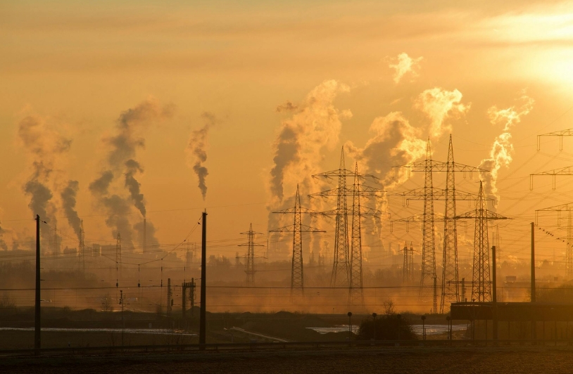 Industrial factory emissions and power lines silhouetted against a hazy sky during sunset, highlighting air pollution and climate-related risks to workers. Caption: Industrial pollution, like that seen here, highlights the environmental hazards worsened by climate change, posing unique threats to workers in vulnerable areas like the Tennessee plastics factory tragedy.