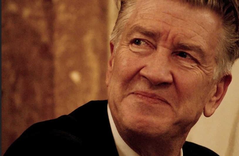 Portrait of David Lynch, the acclaimed filmmaker, smiling subtly while wearing a suit and tie,