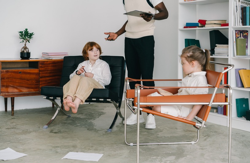 Two children sitting in modern chairs while an adult holding a clipboard gestures nearby, with a bookshelf and wooden sideboard in the background