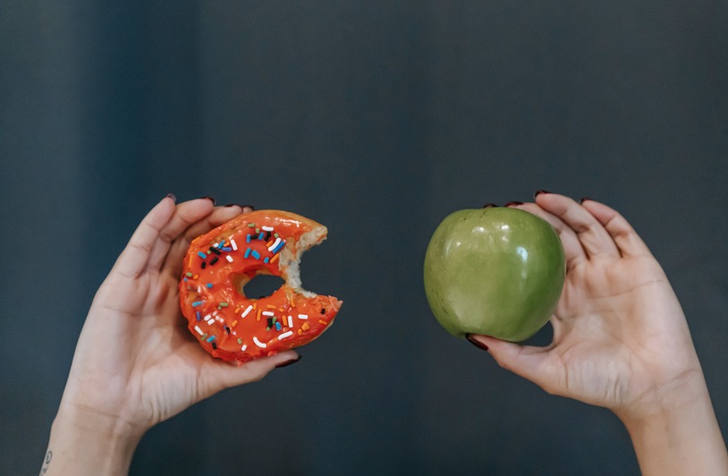 Behavioral Economics of Temptation Crop unrecognizable young female demonstrating bitten high calorie doughnut and healthy green apple against gray background