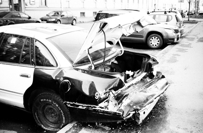 Black and white photo of a severely damaged car with crumpled hood and front end, parked on a street with other vehicles nearby