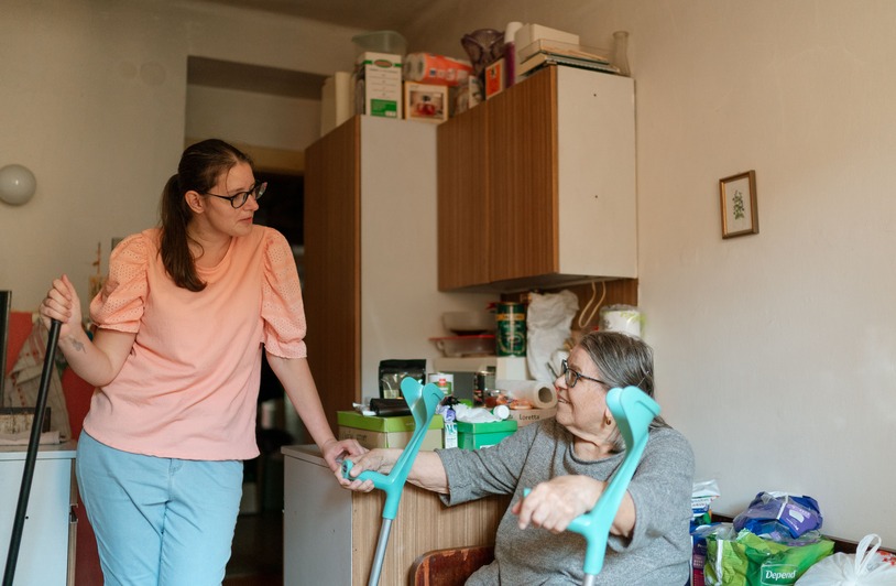 A caregiver engaging with a senior using crutches in a cozy Prague home environment.