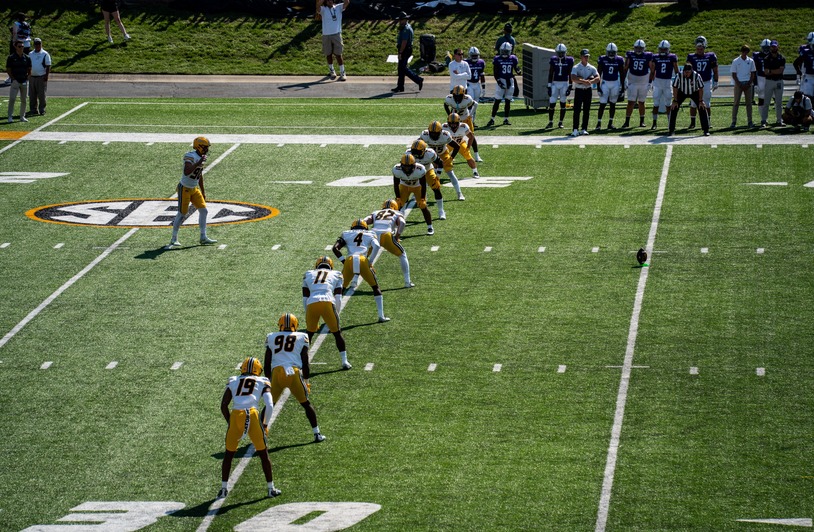 Football players in white and gold uniforms lined up on a green field facing a team in dark blue jerseys near the sideline