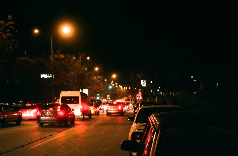 Nighttime street with multiple cars showing red brake lights, a white van, and orange street lamps illuminating a busy road lined with trees