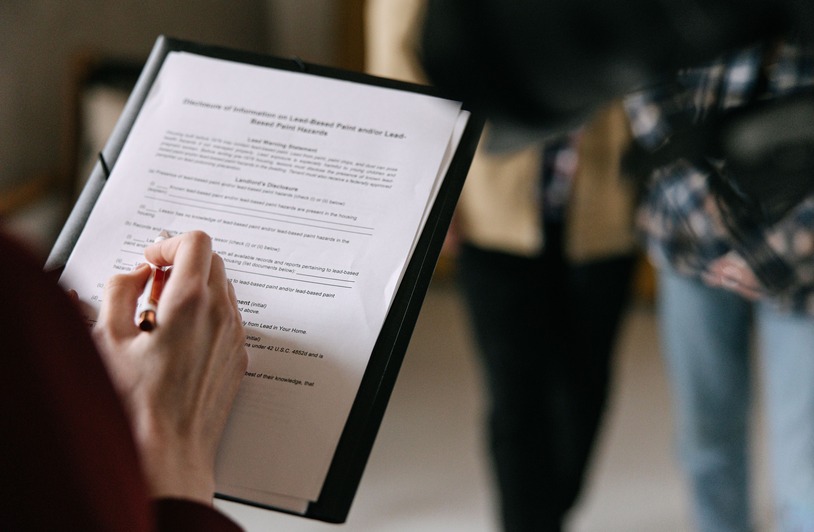 Trusts Explained: Why Estate Planning Today Can Protect Your Family Hand holding a pen over a multi-page document on a black clipboard, with a blurred person standing nearby in jeans and a plaid shirt