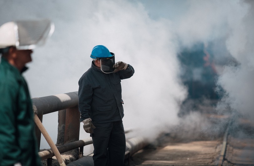 Why Industrial Air Quality Matters to Businesses Worker in blue hard hat and face shield standing amid thick white smoke near metal pipes, second worker in green visible at left