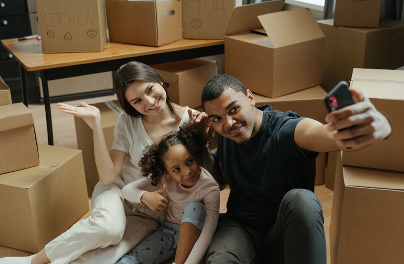 How an International Move Changes What Families Need From a Home Two adults and a young girl sit on a floor taking a selfie, surrounded by stacked cardboard moving boxes