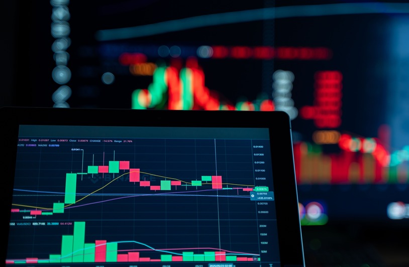 Laptop screen displaying a candlestick trading chart with red and green bars, blurred monitors with colorful charts in background