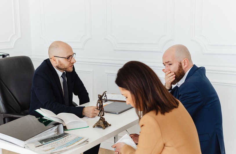What Should You Bring to an Injury Lawyer? Three people seated at a white desk with open books, a laptop, and a small Lady Justice statue