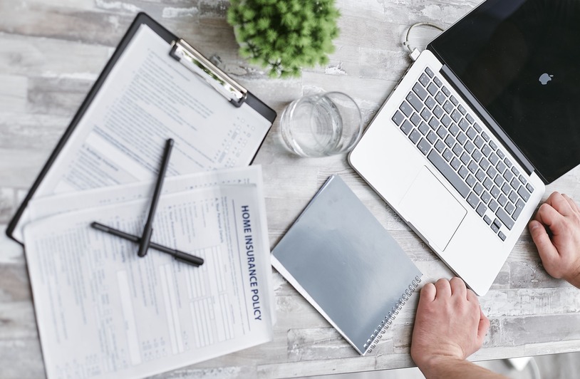 How Continuing Education Helps Mortgage Professionals Stay Competitive Overhead view of a desk with a laptop, clipboard holding papers labeled Home Insurance Policy, two pens, a notebook, glass of water, small plant, and a person's hands