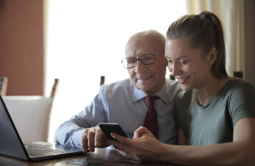 Modern Tools That Improve Safety and Accountability in Elder Care Settings Positive senior man in formal shirt and eyeglasses and smiling young granddaughter sharing mobile phone while sitting near laptop at table