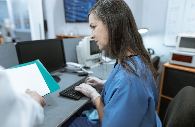 Woman in blue scrubs and gloves typing at a desktop keyboard while another person holds a teal folder nearby