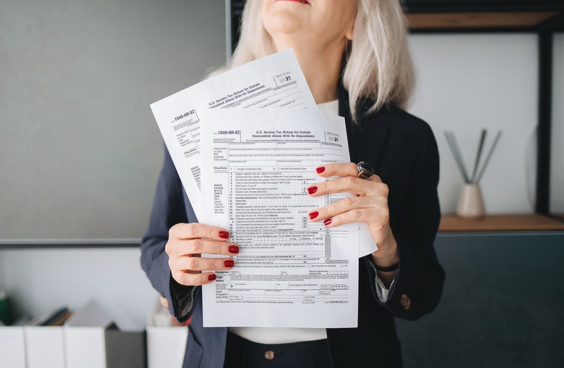8 Tax Strategies Real Estate Investors Woman with gray hair and red nails holding multiple IRS 1040-NR-EZ tax forms, wearing a dark blazer indoors