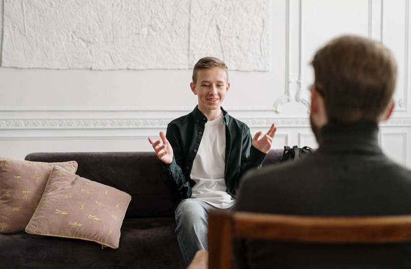 Teen in green shirt gesturing with hands while seated on dark sofa, facing an adult seen from behind in a chair