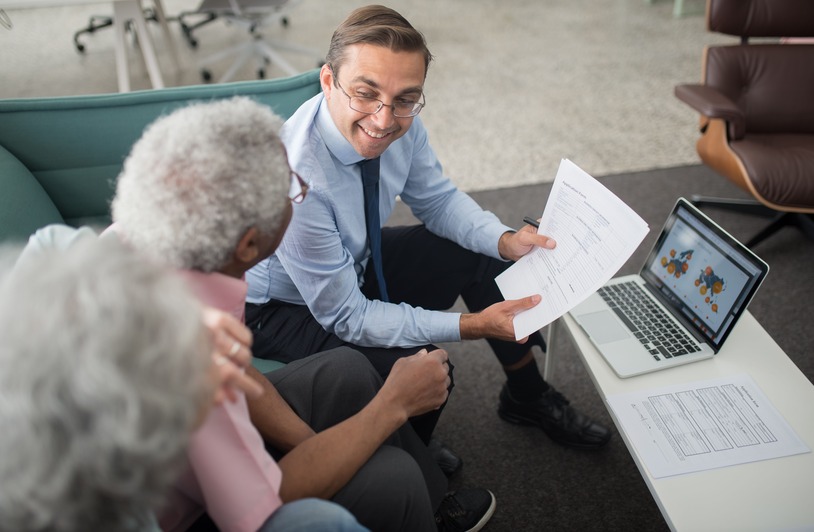 Man in blue shirt and tie holding papers, smiling at two older adults seated on a teal sofa, laptop and documents on a low table nearby