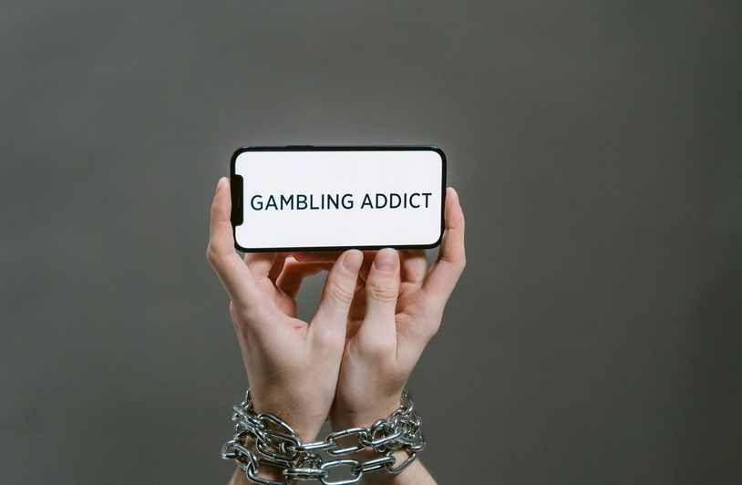 Chained hands hold a phone displaying the text 'Gambling Addict', highlighting addiction.