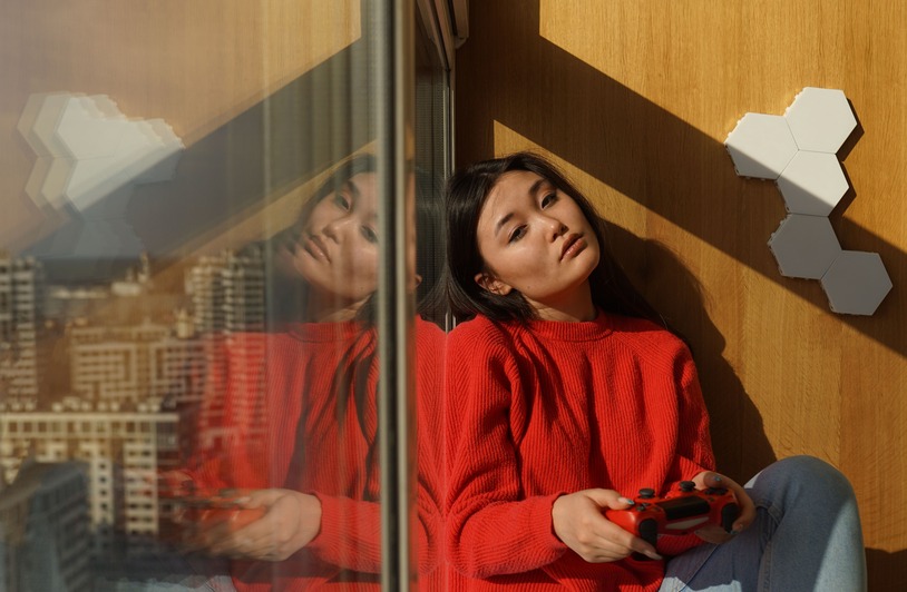 Does Solo Gaming Still Have its Place? 5 Reasons it Does Woman in red sweater and jeans leaning against a glass window holding a red game controller, with hexagonal wall panels and a city skyline reflection visible