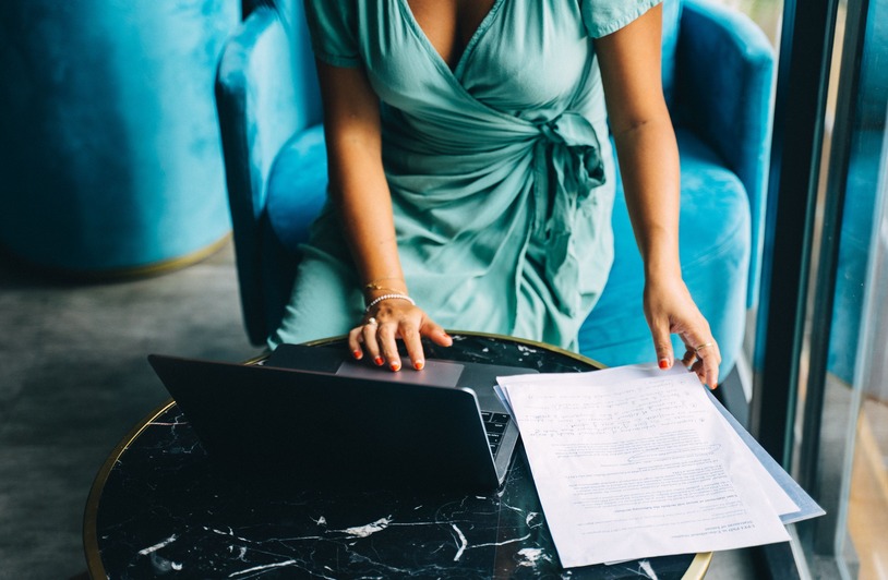 Woman in teal wrap dress leaning over a marble table with a laptop and printed documents, blue velvet chair behind her