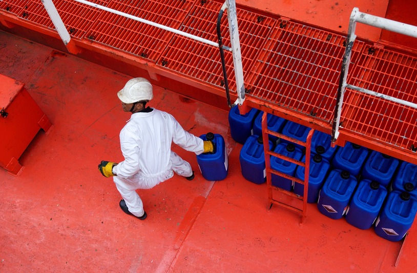 Texas Chemical Spill Could Bring $3.5 Million in OSHA Fines Person in white coveralls, hard hat, and yellow gloves carrying a blue plastic container on a red industrial deck with more blue containers nearby