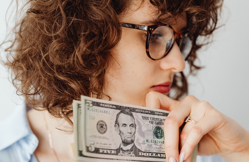 The Side-Income Obsession Changing How Americans Think About Work Close-up of a woman with glasses holding US dollar bills. Thoughtful expression.