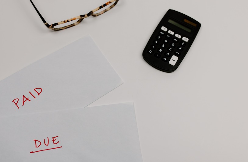 Understanding the True Cost of Business Debt Two white envelopes labeled PAID and DUE in red handwriting beside a black calculator and tortoiseshell glasses on a white surface