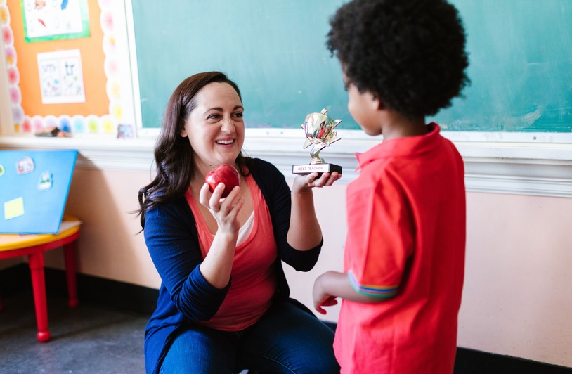 A teacher holding an apple and a student holding a trophy in a classroom setting.