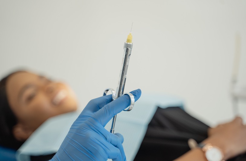 Injector Training: What Makes a Qualified Aesthetic Injector? Skills Blue-gloved hand holding a metal syringe with a smiling patient lying in a chair behind