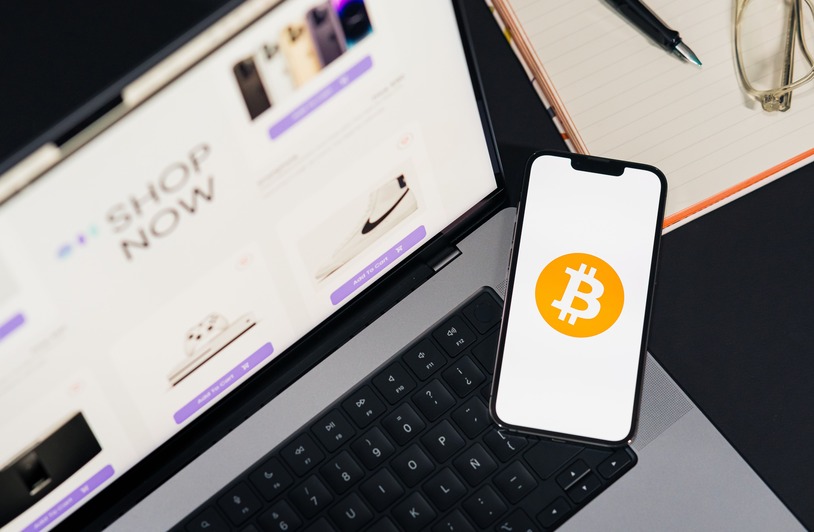 Smartphone displaying Bitcoin logo resting on a laptop keyboard showing a Shop Now webpage, with a notepad and glasses nearby