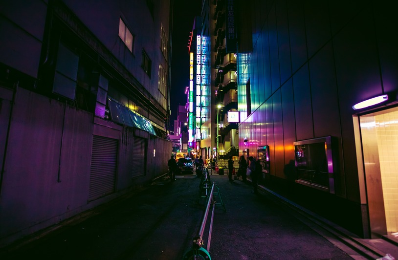 Solo Trip to Tokyo: A Modern Urban Backpacker’s Guide Narrow dark alley at night with neon signs glowing in purple, green, and yellow, flanked by tall buildings and a few distant pedestrians