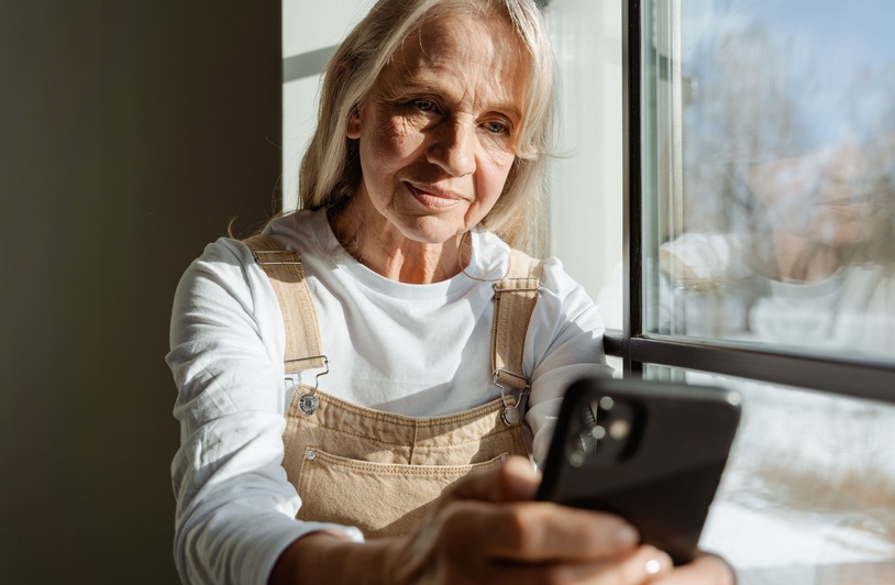 Long-Distance Caregiving: How to Support an Aging Parent From Far Away A senior woman enjoys leisure time browsing on her smartphone by a sunlit window.
