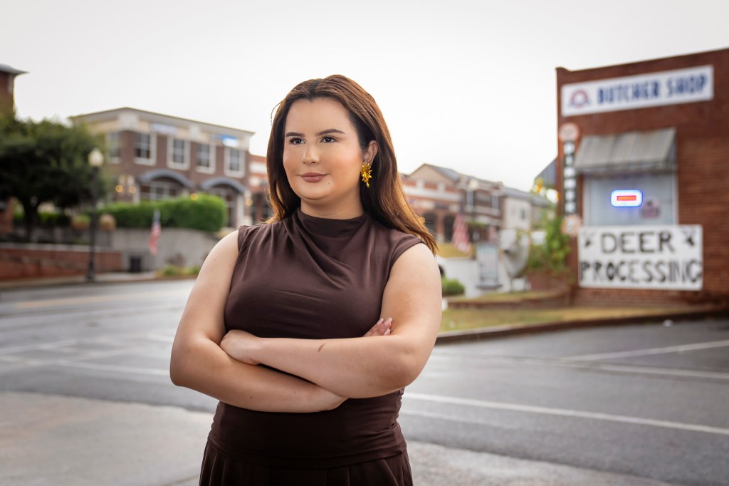 Portrait of Bautista standing arms crossed in the middle of an empty rural street with a building behind her that says "Deer processing.